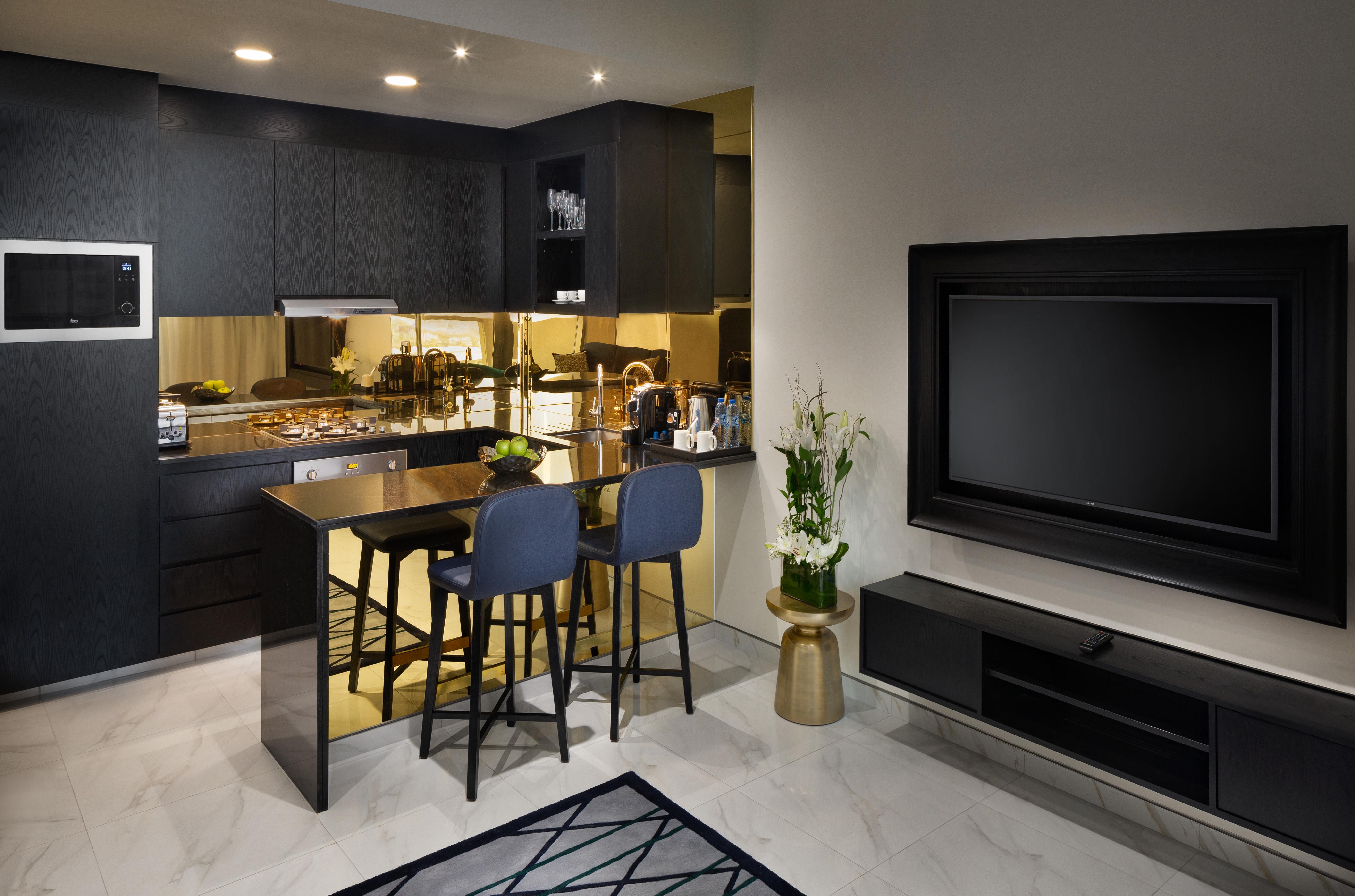 Millennium Atria Business Bay Aparthotel Дубай Екстериор снимка The photo depicts a modern kitchen and living area. The kitchen features dark cabinetry with a clean design, showcasing various appliances, including a stovetop and possibly an oven. There is a kitchen island with two stools, suggesting a space for c