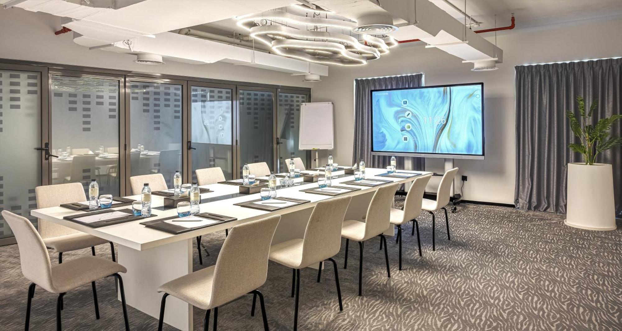 Millennium Atria Business Bay Aparthotel Дубай Екстериор снимка The photo shows a modern conference room set up for a meeting. There is a long table with several chairs arranged around it, each with a notepad and pen. A large screen displays a graphic or presentation, and there is a whiteboard in the corner. The 