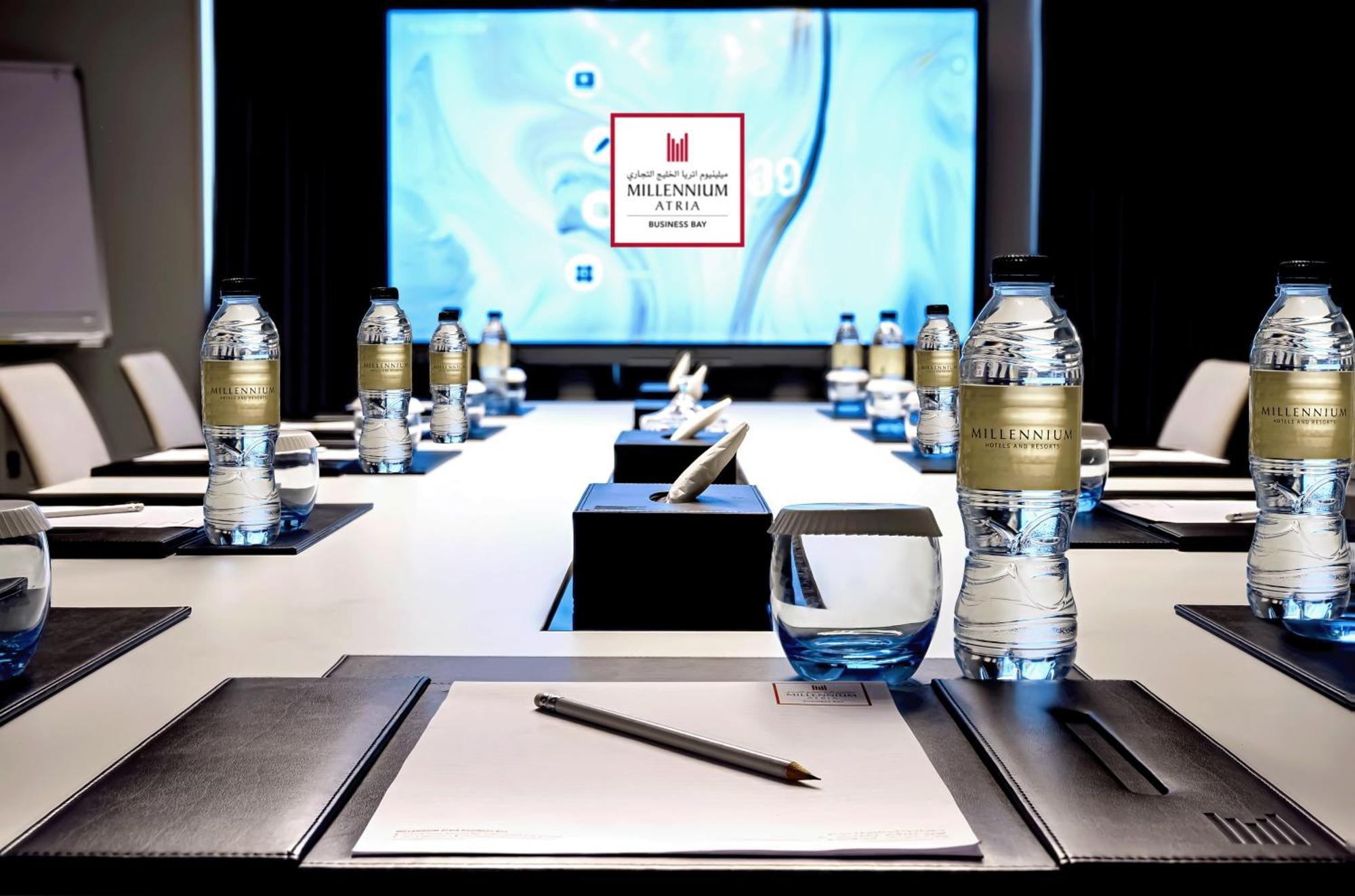 Millennium Atria Business Bay Aparthotel Дубай Екстериор снимка The photo shows a modern conference room setup. A long table is laid out with neatly arranged stationery, including notebooks and pens, at each seat. Several bottles of water are placed on the table. In the background, a large screen is displayed, li