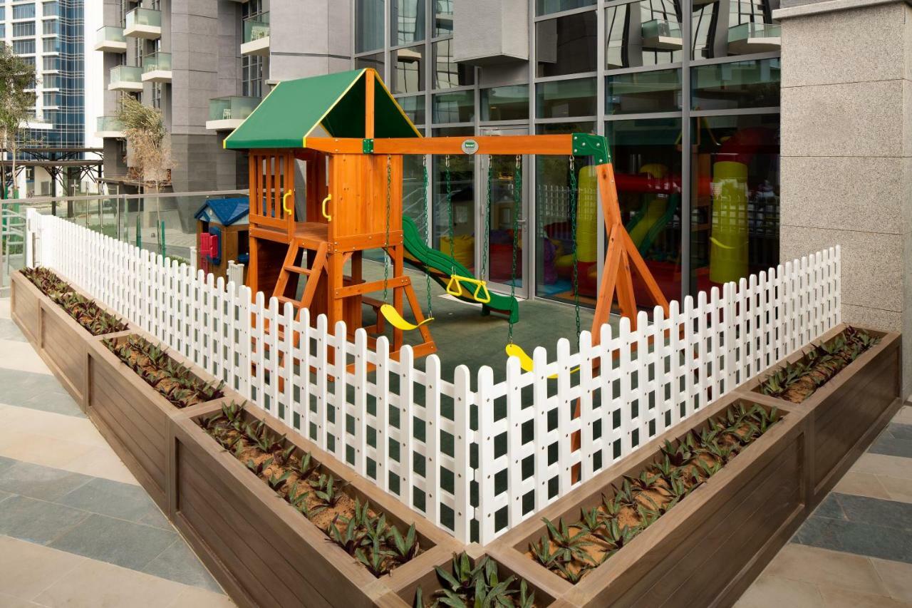 Millennium Atria Business Bay Aparthotel Дубай Екстериор снимка The image shows a children's playground area, which is enclosed by a white picket fence. The playground features a wooden play structure with a slide, swings, and climbing elements. Surrounding the play area are planters with greenery, and the backdr