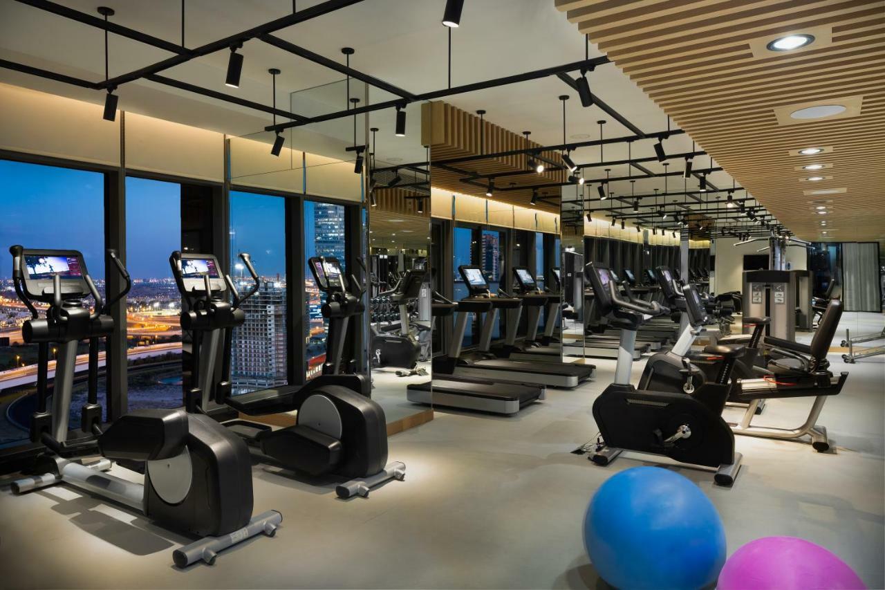 Millennium Atria Business Bay Aparthotel Дубай Екстериор снимка The photo shows a modern gym with various types of exercise equipment available. There are several treadmills and elliptical machines arranged in rows, with large windows in the background providing a view of the city. The lighting is bright and cont