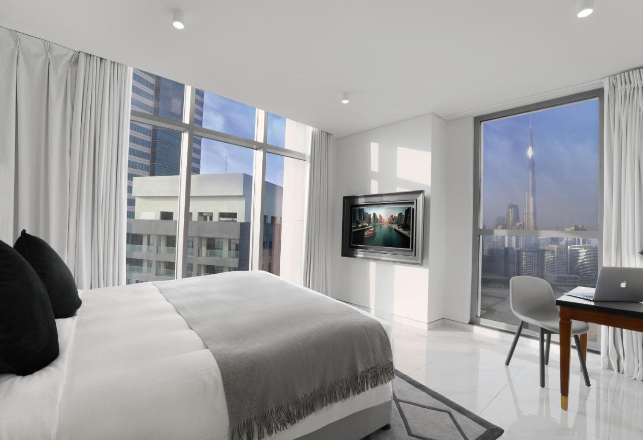 Millennium Atria Business Bay Aparthotel Дубай Екстериор снимка The photo depicts a modern hotel room that features a large bed with a gray throw blanket. There are large windows allowing natural light to fill the space, showcasing a city skyline view. A flat-screen television is mounted on the wall opposite the 